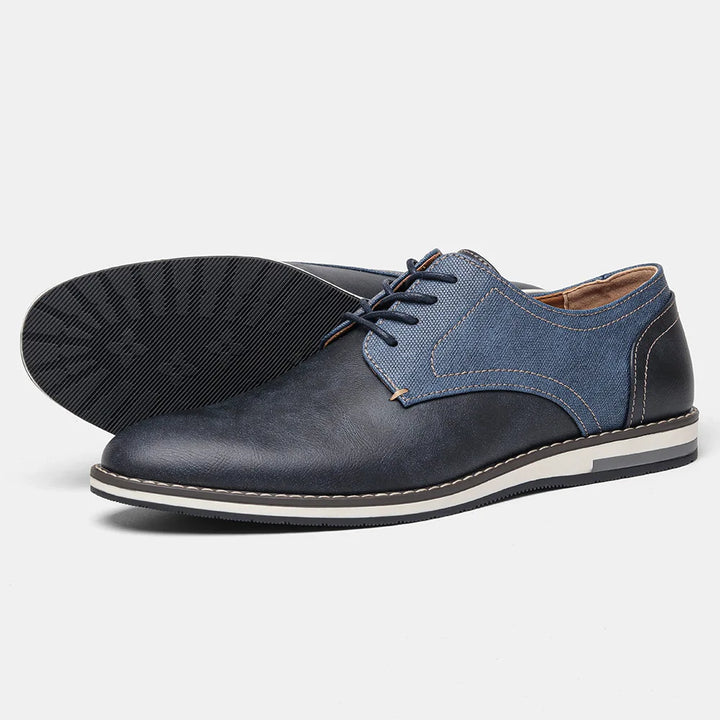 A pair of stylish mens lace-up patchwork oxfords in dark blue with denim-textured panels, white stitching, and a cushioned sole. One shoe is upright while the other is laid on its side to show the sole.