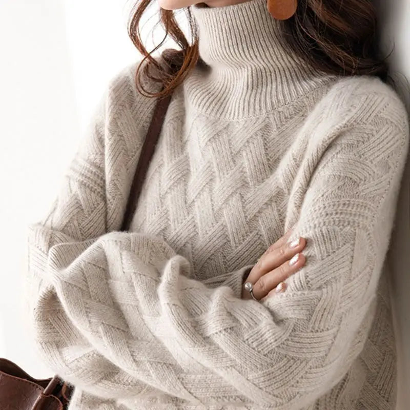 A person wearing a cozy, cream-colored, cashmere blend cable-knit turtleneck sweater with a zigzag knit pattern. They have long brown hair, are carrying a brown bag over their shoulder, and their arms are folded.