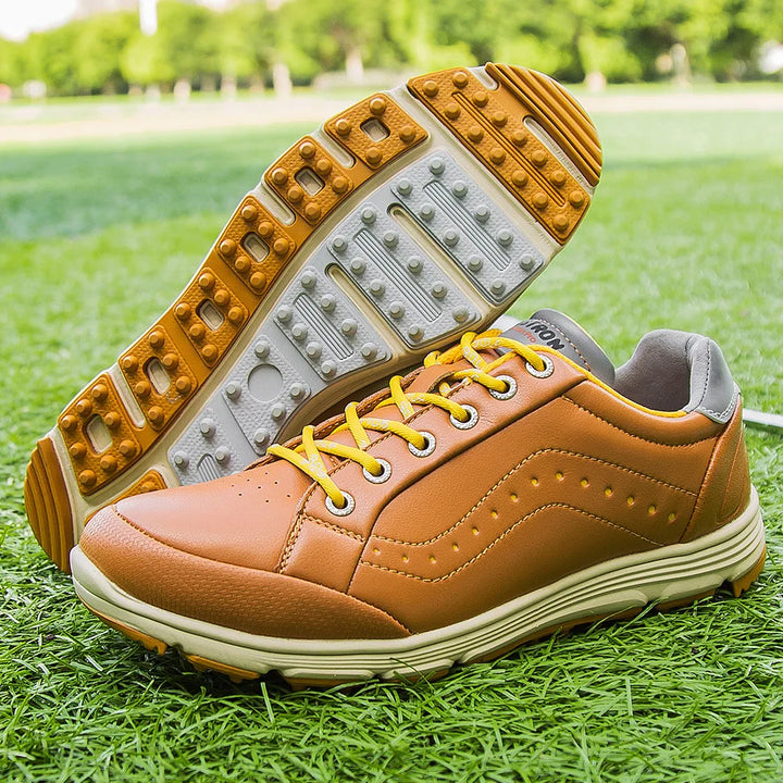 A pair of brown golf footwear with yellow laces, featuring white and orange rubber soles and advanced cleat technology, displayed on green grass with a blurred outdoor background.
