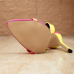 A close-up of a single high-heeled shoe with stiletto heels lying on its side, showing the beige sole, bright yellow upper, and pink trim, set against a tan textured background and floor.