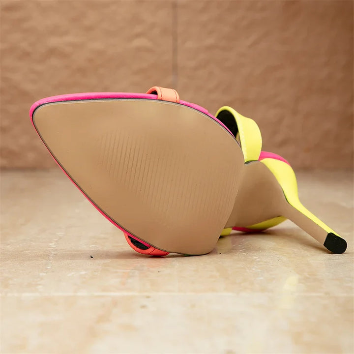 A close-up of a single high-heeled shoe with stiletto heels lying on its side, showing the beige sole, bright yellow upper, and pink trim, set against a tan textured background and floor.