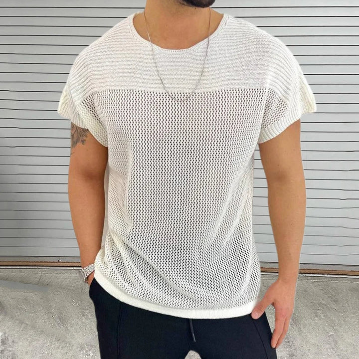 A man in a loose, white mesh knit tee with a breathable design, black pants, a silver chain necklace, and a watch stands in front of a gray horizontal metal shutter—perfect streetwear style.