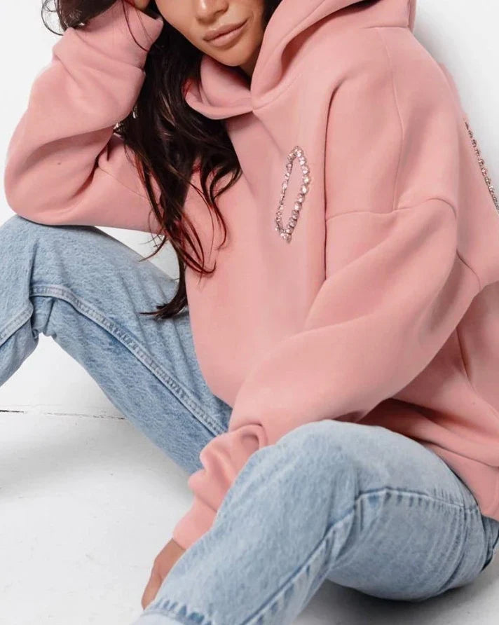 A woman sits on the floor wearing an oversized pink hoodie with a heart-shaped design and light blue jeans. Her head is slightly tilted, and she is leaning her arm on her knee.