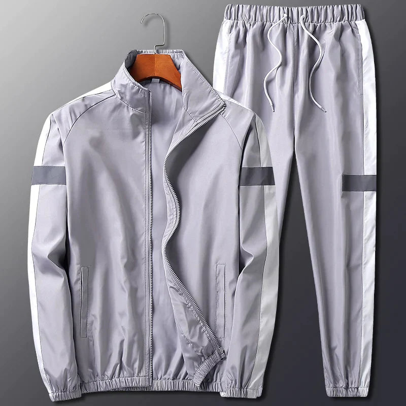 A light gray activewear two-piece set with white side stripes features a zip-up jacket with a high collar and matching pants with an elastic waistband and cuffs. Both pieces have dark gray accent stripes on the sleeves and legs.