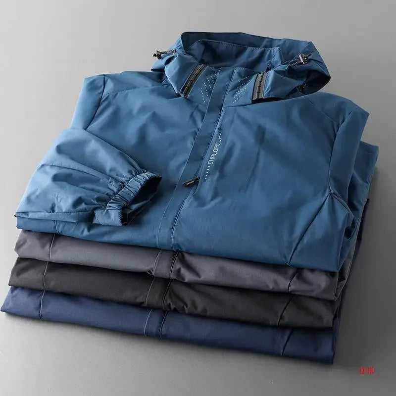 Four folded jackets in varying dark shades, with the top one being a blue, breathable outdoor jacket featuring a zippered front and a hood, neatly stacked on a gray surface.