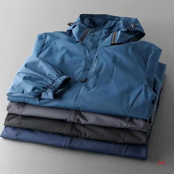 Four folded jackets in varying dark shades, with the top one being a blue, breathable outdoor jacket featuring a zippered front and a hood, neatly stacked on a gray surface.