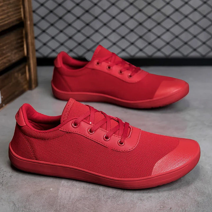 A pair of bright red men's canvas sneakers with laces, placed on a gray floor. One shoe stands upright, while the other is set behind it and tilted to display the outer side. A wire mesh wall serves as the background.