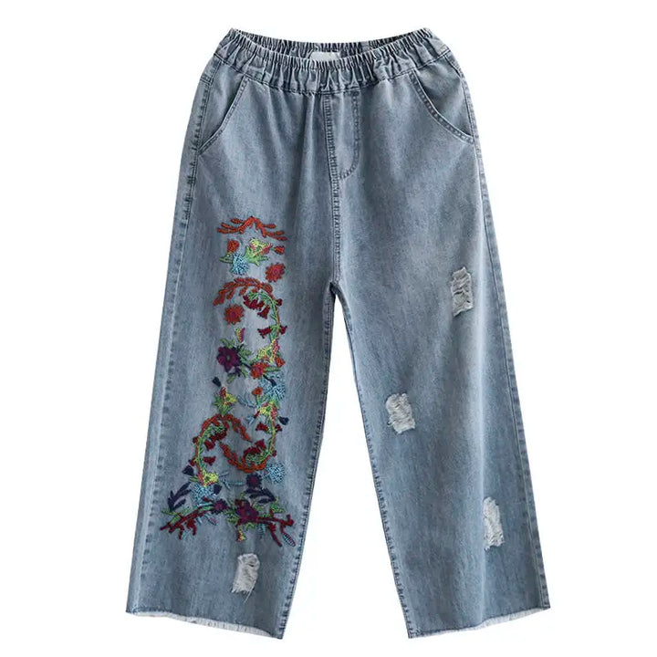Light blue loose-fitting denim wide-leg jeans with an elastic waistband, front pockets, distressed patches, and a bold retro embroidery floral design running vertically down one leg.
