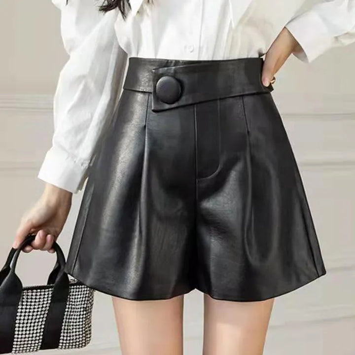 A woman wears high waist black faux calfskin leather shorts with a striking button detail, paired with a white blouse. She carries a small black and white checkered handbag.