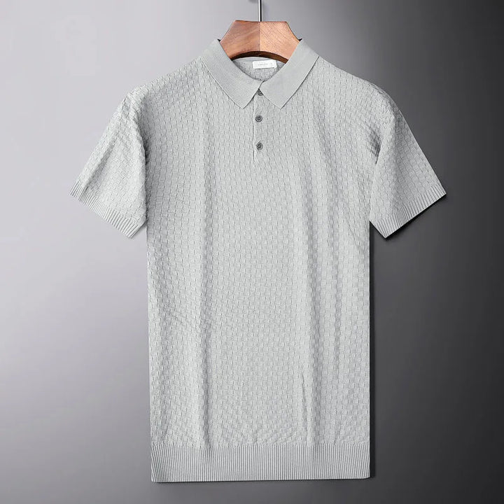 A light gray short-sleeve knit polo shirt with a subtle waffle jacquard pattern hangs on a wooden hanger against a gray background. The shirt features a collar and a three-button placket.