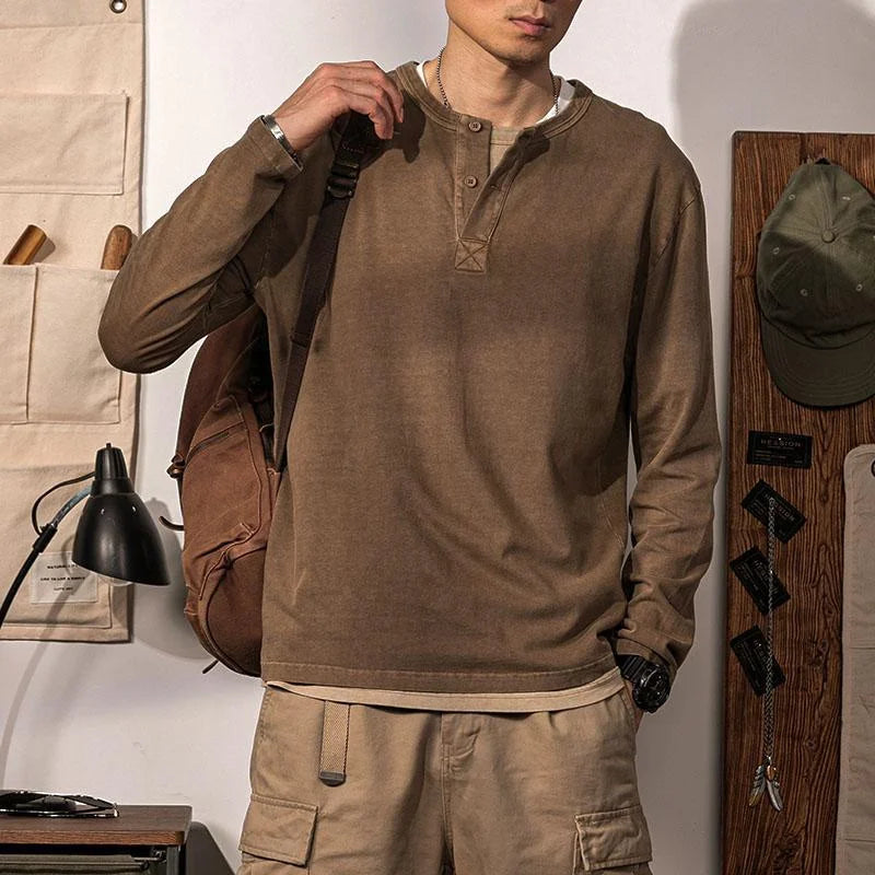 A person stands indoors wearing a brown pure cotton Henley shirt and beige cargo shorts, with a brown backpack over one shoulder. The room has a desk lamp, organizer, hats, and accessories on the wall.