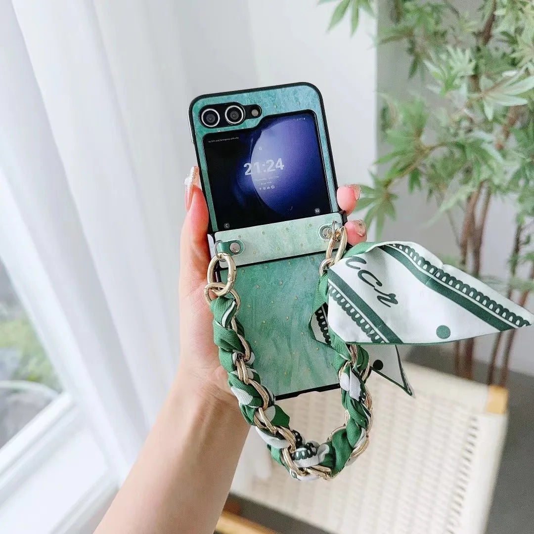 A hand holds a green foldable smartphone with a matching marbled case featuring shockproof protection, a gold and green braided chain strap, and a green-and-white scarf attached. In the background, there’s a chair and a leafy potted plant.