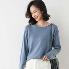 A woman with shoulder-length dark hair smiles while looking down. She is wearing a relaxed fit, light blue long-sleeve top, white pants, and carrying a dark shoulder bag. The background is plain and light-colored.
