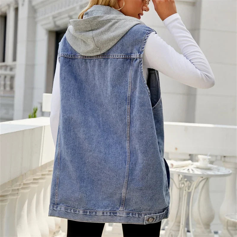 A person stands on a balcony facing away, wearing a light gray hooded sweatshirt under a sleeveless denim vest made from pre shrunk denim and black pants. The background features white architectural details and outdoor furniture.