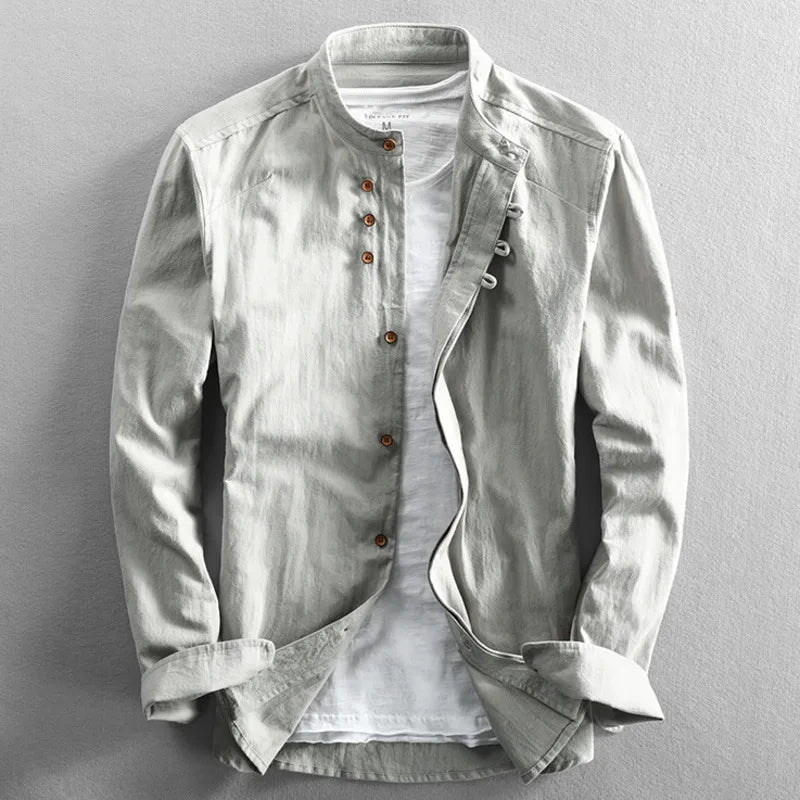 A light gray, long-sleeved, Japanese inspired overshirt with a mandarin collar and brown buttons is displayed over a plain white t-shirt on a flat, neutral background. The 100% cotton jacket is partially unbuttoned and the sleeves are slightly folded.