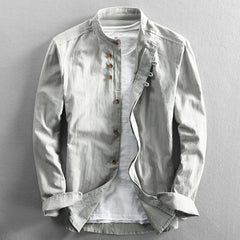 A light gray, long-sleeved, Japanese inspired overshirt with a mandarin collar and brown buttons is displayed over a plain white t-shirt on a flat, neutral background. The 100% cotton jacket is partially unbuttoned and the sleeves are slightly folded.