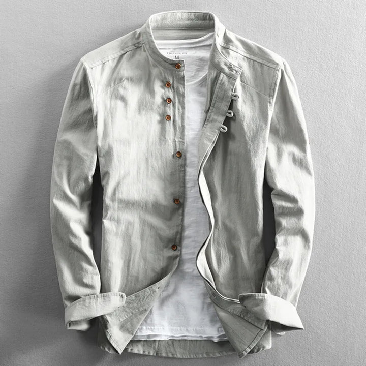 A light gray, long-sleeved, Japanese inspired overshirt with a mandarin collar and brown buttons is displayed over a plain white t-shirt on a flat, neutral background. The 100% cotton jacket is partially unbuttoned and the sleeves are slightly folded.