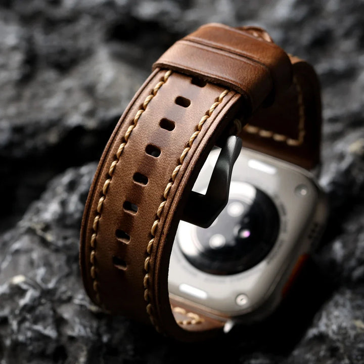A close-up of a smartwatch with a handcrafted cowhide leather strap featuring light stitching, set against a textured gray rock background. The watch face is partially visible, focusing on the strap and metal buckle.