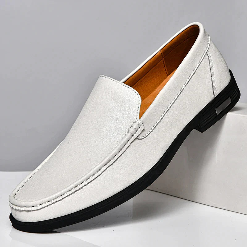 A classic slip-on white leather loafer with a black sole, tan interior, and visible stitching is displayed against a light gray background, its heel propped on a white block. Experience the elegance of genuine leather loafers.