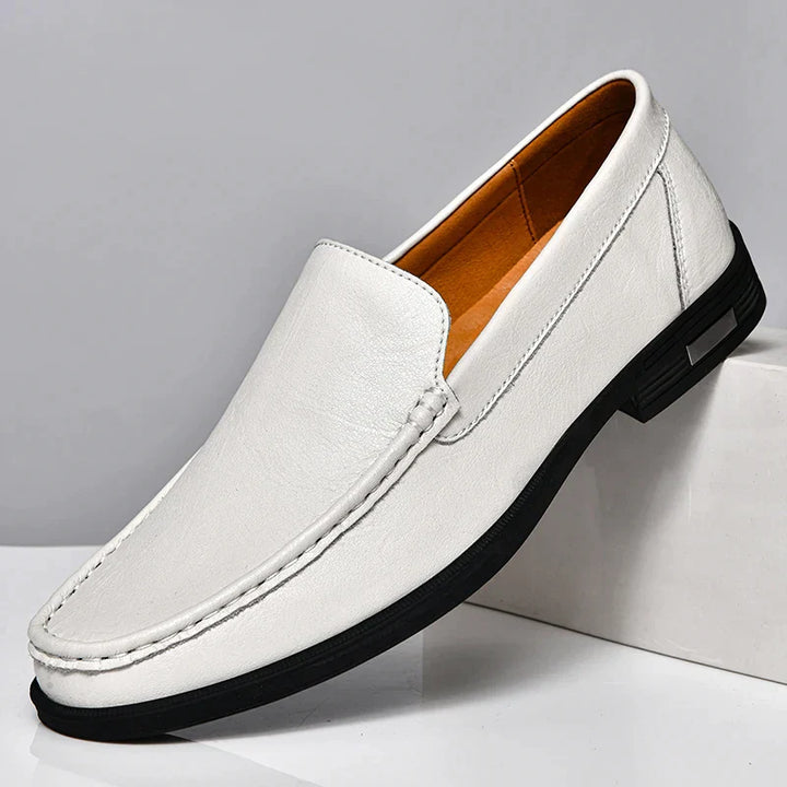 A classic slip-on white leather loafer with a black sole, tan interior, and visible stitching is displayed against a light gray background, its heel propped on a white block. Experience the elegance of genuine leather loafers.