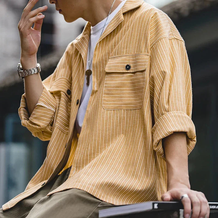 A person wearing a loose, high-quality cotton yellow and white pinstripe shirt with rolled-up sleeves sits casually outdoors. The shirt has front pockets, and the look is completed with a necklace and a wristwatch.