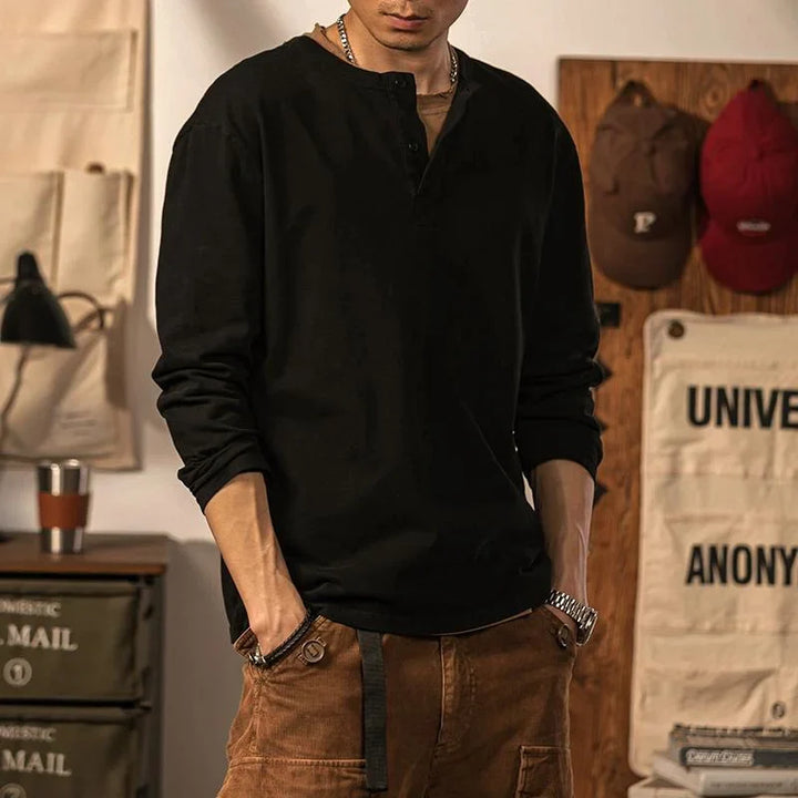 A man wearing a black pure cotton Henley shirt and brown pants stands indoors with his hands in his pockets. The background includes wall decorations, hats, and storage bins. His face is partially out of frame.