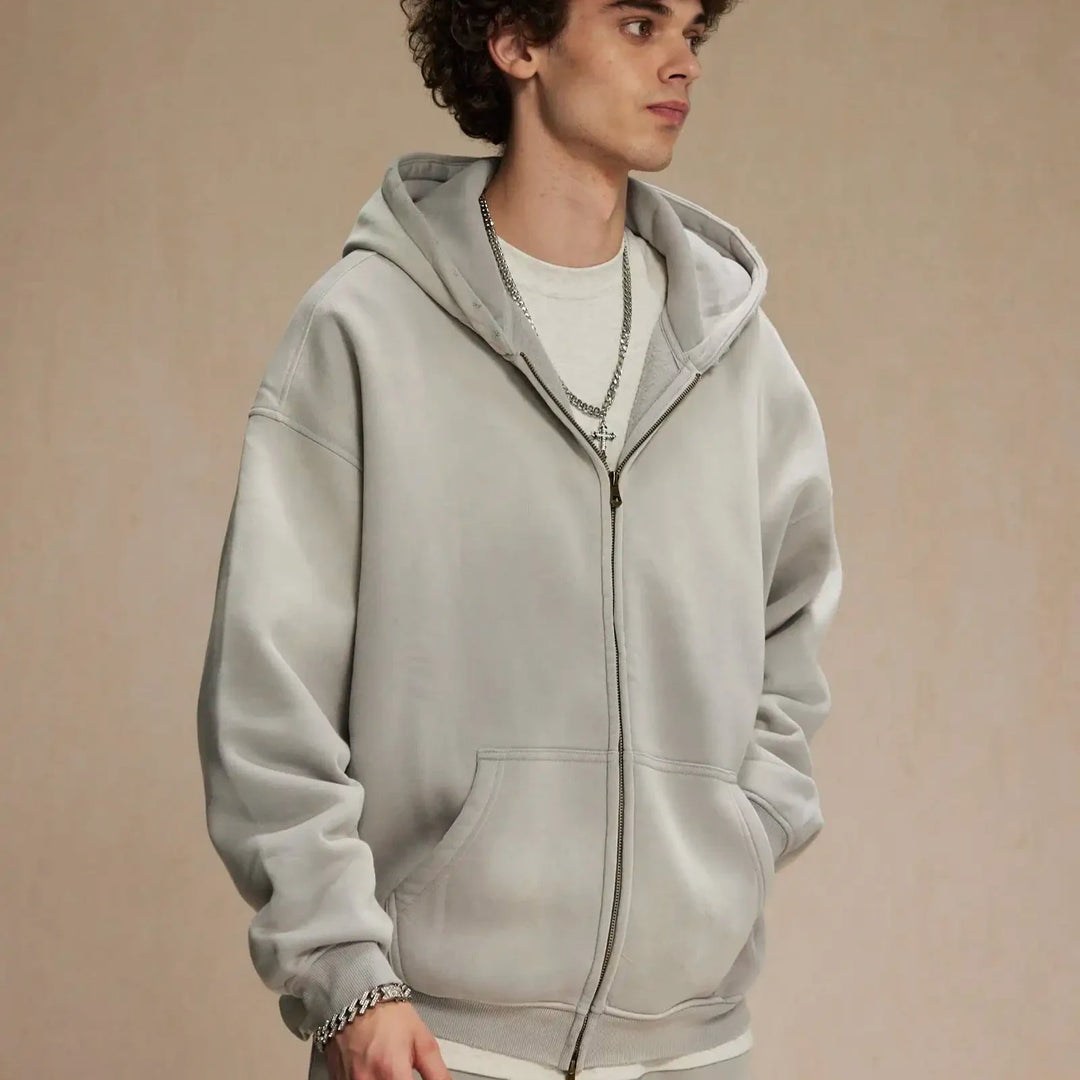 A young person with curly hair wears a light gray zip-up hoodie made of thickened fabric over a white shirt, accessorized with silver chains and a bracelet, standing against a plain beige background.