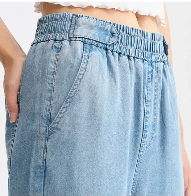 Close-up of a person wearing light blue, high-waisted, wide-leg denim pants with an elastic waistband and side pockets; the person’s hand is in one pocket, and a white cropped top is partially visible.