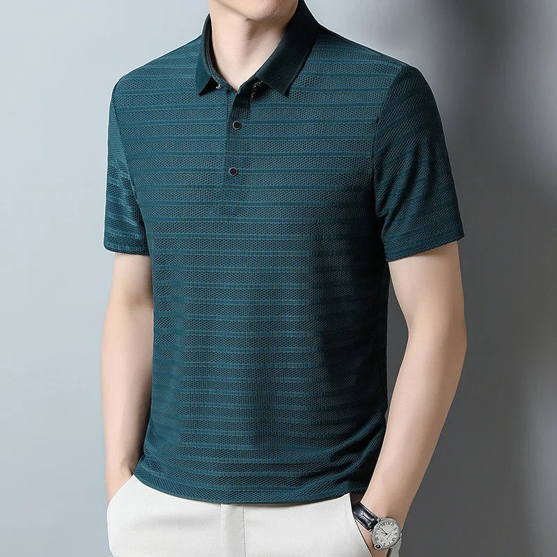 A man wearing a dark green, short-sleeve mens silk polo shirt with a subtle horizontal pattern, paired with light-colored pants and a watch, stands against a plain light gray background.