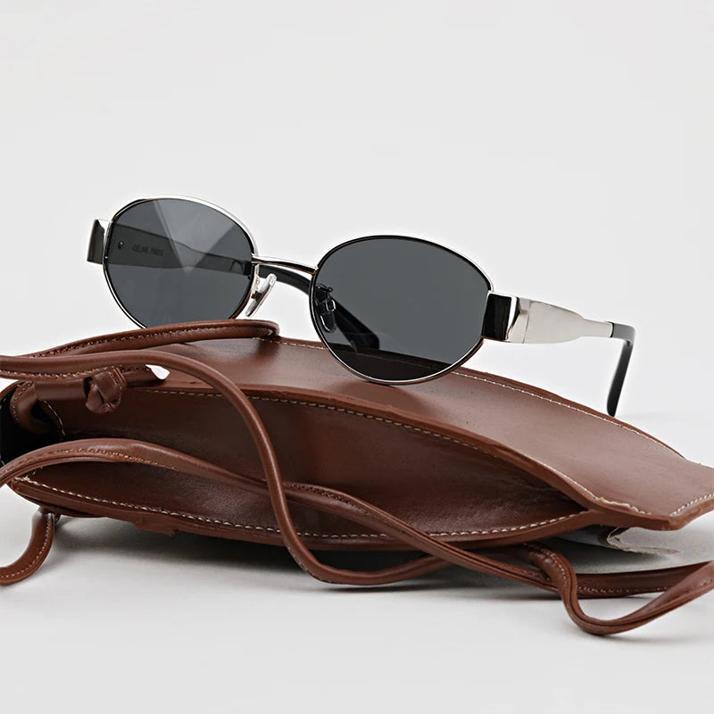 A pair of black oval, vintage-inspired sunglasses with silver frames rests on top of a brown leather pouch with a strap against a plain white background.
