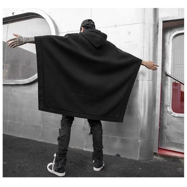 A person wearing a black Techwear hoodie and black jeans stands with arms outstretched, facing a metallic wall with a window and a door. The scene appears to be urban and industrial.