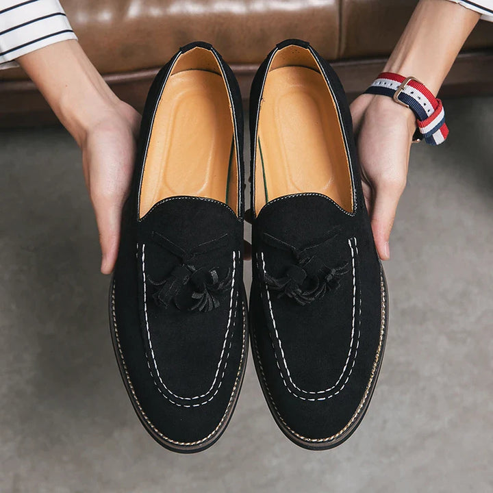A person holds a pair of black suede tassel loafers with white stitching, tan insoles, and cushioned footbed, above a gray floor.