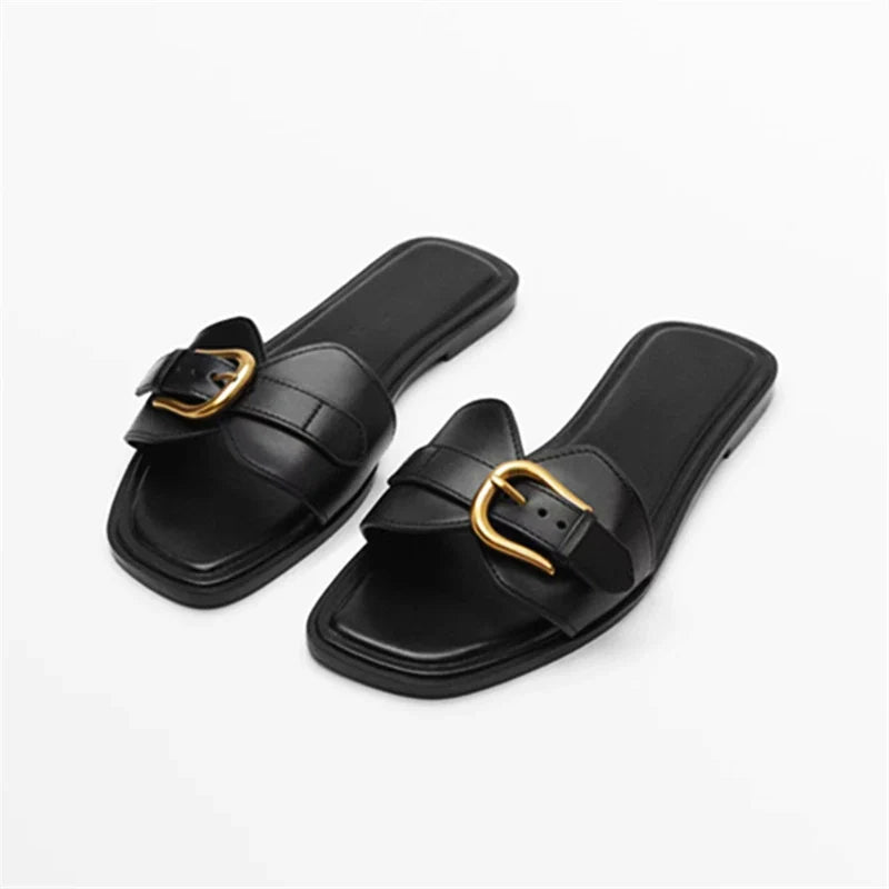A pair of black, flat leather slides with square toes, wide straps, and a gold buckle, displayed on a white background.