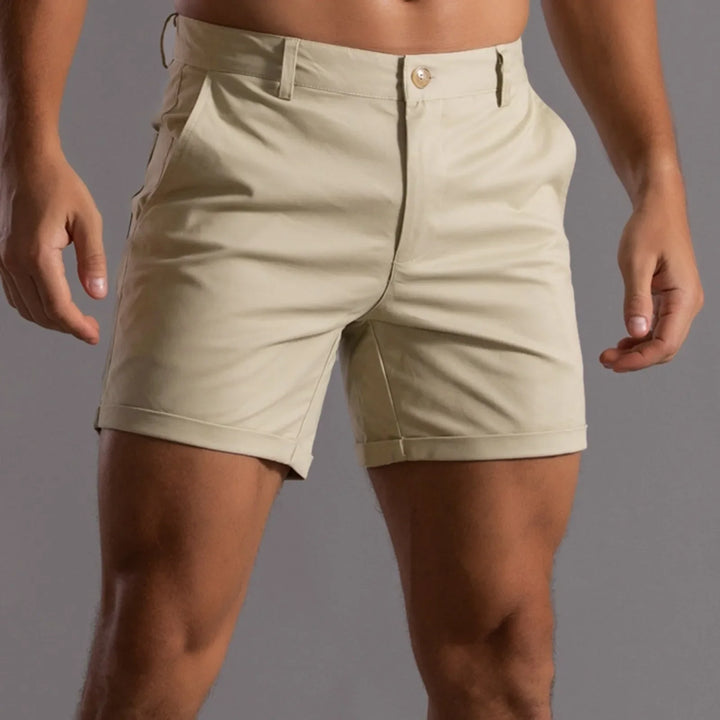 A person wearing light beige shorts with a semi elastic waistband and tailored fit, standing with hands slightly out to the sides against a plain gray background. The shorts have a button and zipper closure plus side pockets.