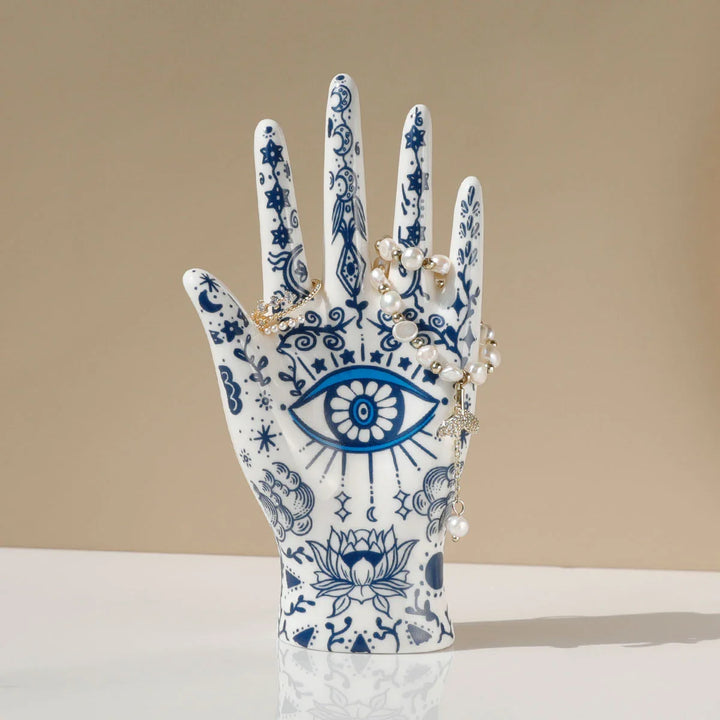 A white ceramic jewelry stand, adorned with blue intricate patterns including the iconic evil eye decor and floral designs, holds a string of pearl beads against a beige background—perfect for boho-chic home décor.