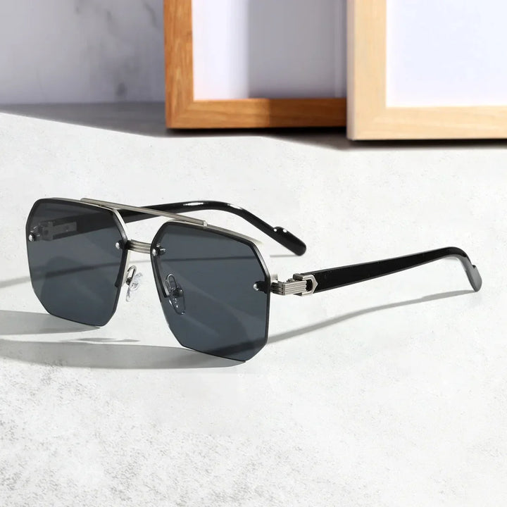 A pair of black-rimmed, geometric metal frame sunglasses with dark UV400 protective lenses rests on a light-colored surface. The frames have a modern, angular design, and the background features blurred picture frames.