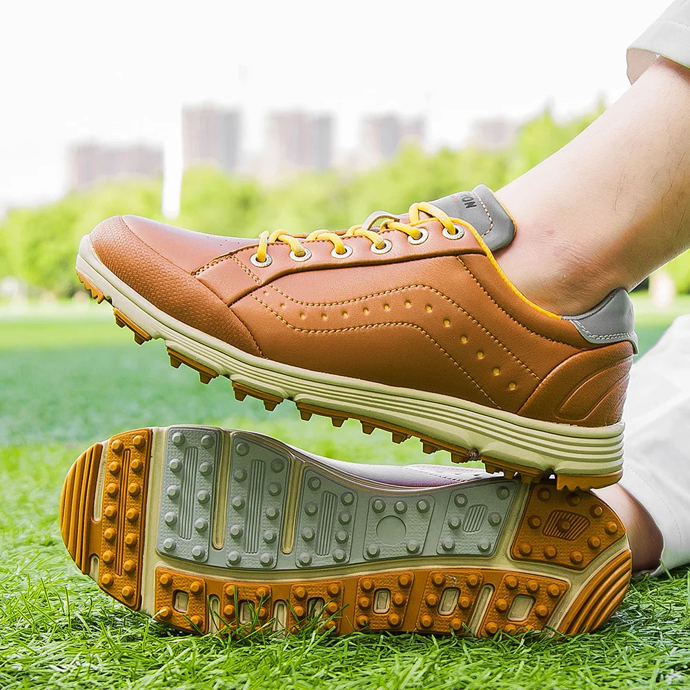 A person wearing brown and yellow golf footwear with advanced cleat technology is sitting on grass, with buildings and trees blurred in the background.