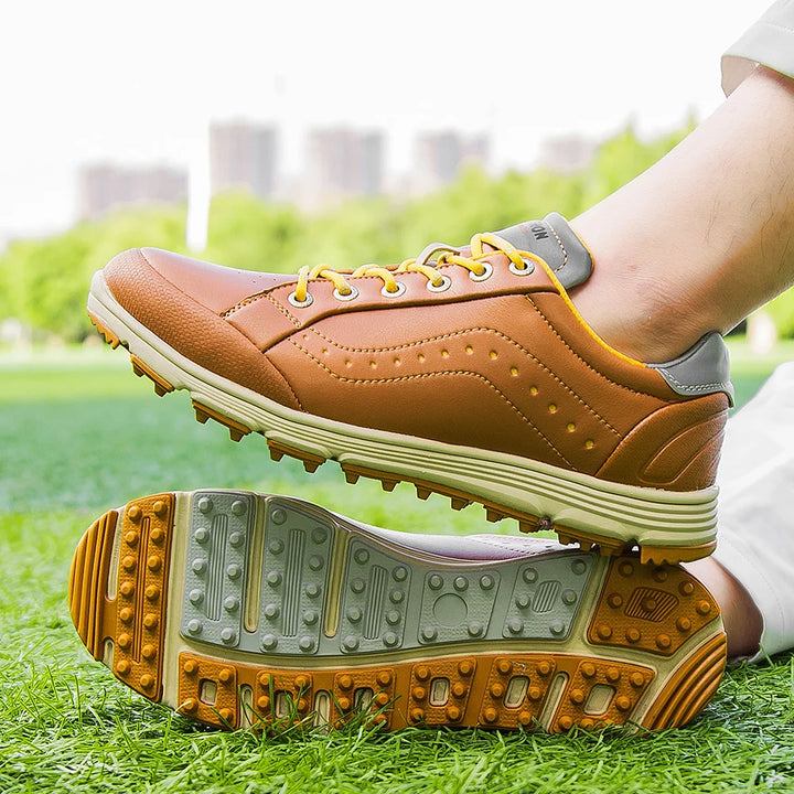 A person wearing brown and yellow golf footwear with advanced cleat technology is sitting on grass, with buildings and trees blurred in the background.