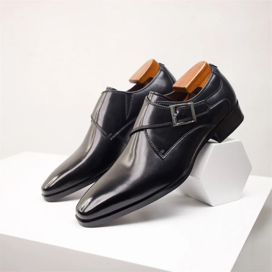 A pair of shiny black Italian leather monk strap shoes with a side buckle and wooden shoe trees, displayed on a white geometric pedestal against a plain background.