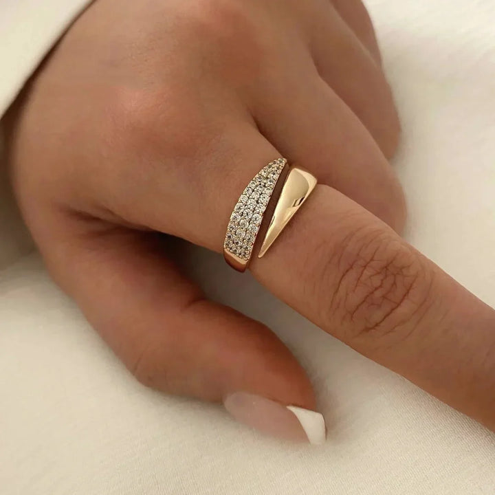 A hand with a light skin tone wears a two-part gold ring featuring a twisted design—one side with a polished finish, the other encrusted with pavé diamonds—on the index finger. The nails are manicured with a white tip.