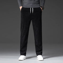 A person wearing a gray sweater, black men's smart casual pants with a zipper fly, and white sneakers stands with hands in pockets against a plain, dark background. Only the body from shoulders down is visible.