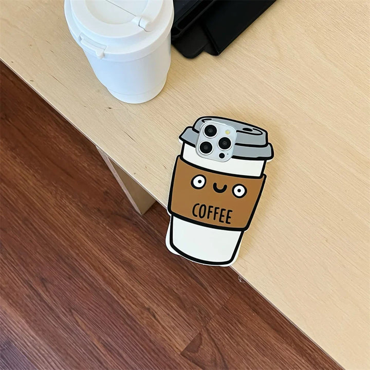 A phone with a 3D coffee cup soft silicone case, featuring a smiling face and the word COFFEE, rests on the edge of a wooden desk near a white reusable cup with lid. The desk stands above a brown wooden floor.