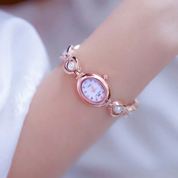 A delicate wristwatch with a rose gold band and small decorative pearl accents is worn on a persons wrist. The oval-shaped watch face features a white background, simple hour markings, and reliable precision quartz movement.