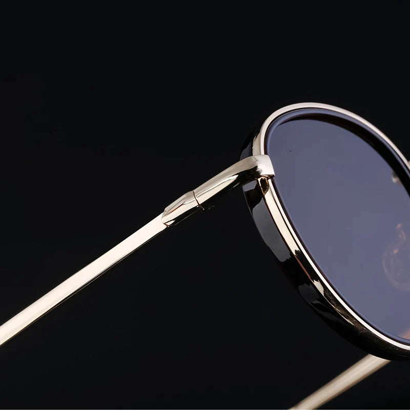Close-up of rounded sunglasses with thin, gold-colored metal arms and dark tinted, scratch resistant lenses featuring UV400 protection, set against a black background.