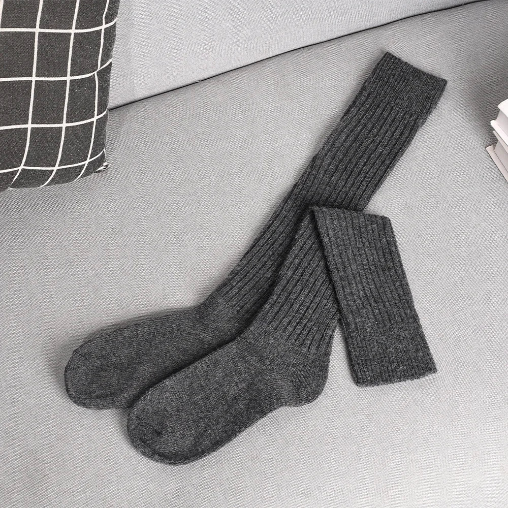 A pair of long, dark gray ribbed socks made from a cashmere cotton blend is laid out on a light gray couch next to a black and white checkered pillow and a small stack of white papers.