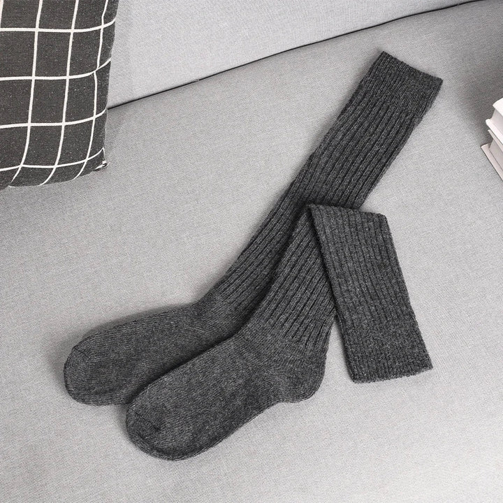 A pair of long, dark gray ribbed socks made from a cashmere cotton blend is laid out on a light gray couch next to a black and white checkered pillow and a small stack of white papers.