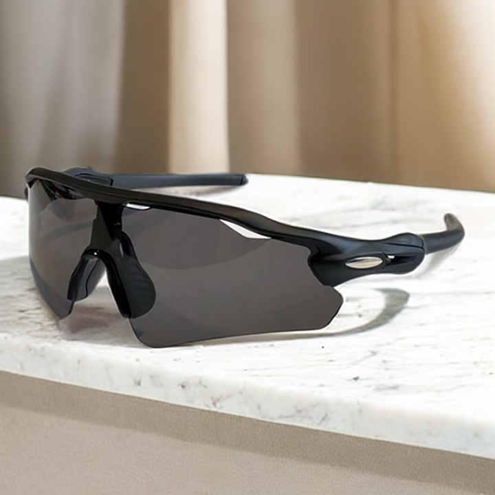 A pair of black, wraparound Polarized Cycling Sunglasses with dark lenses rests on a white marble surface. Beige curtains are visible in the blurred background.