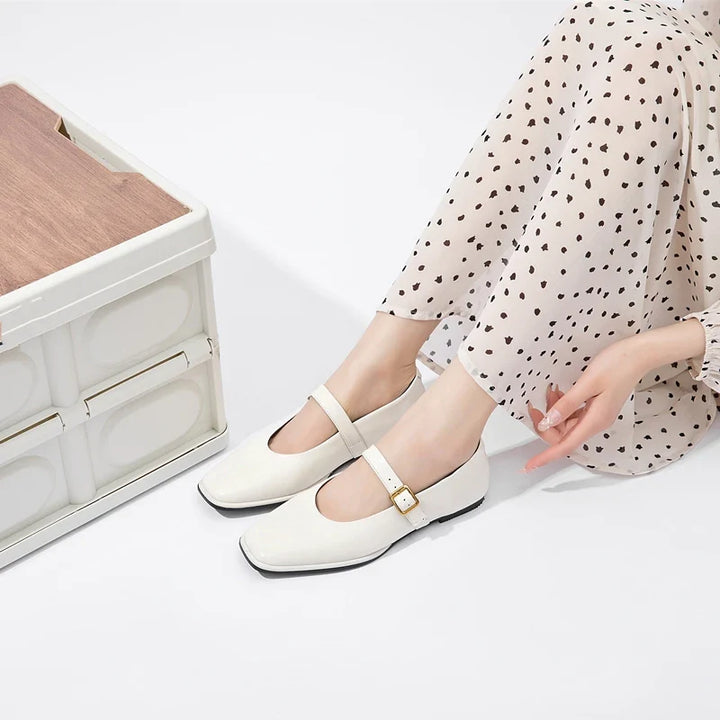 A person wearing cream-colored Augusta Leather Ballet Flats and cream pants with black polka dots sits on the floor next to a white and wooden storage box, with one hand resting on the ground.