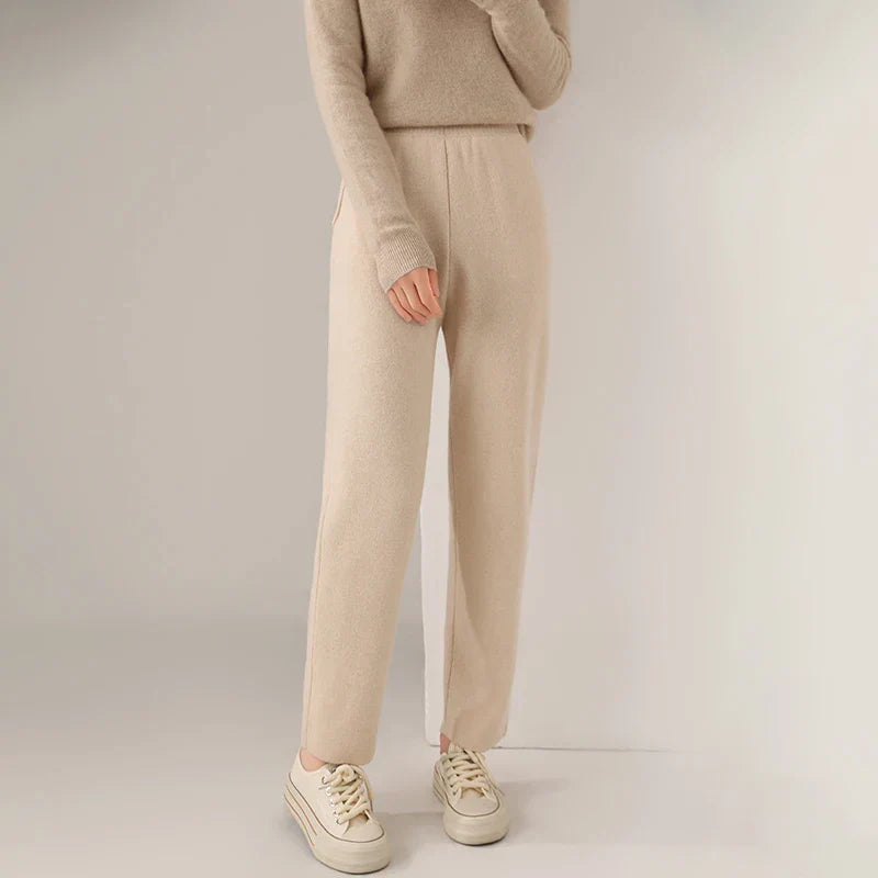 A person wearing a beige long-sleeve sweater, matching beige slim tailored leg pants, and cream-colored sneakers stands against a neutral background. Their head is not visible in the image.