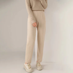 A person wearing a beige long-sleeve sweater, matching beige slim tailored leg pants, and cream-colored sneakers stands against a neutral background. Their head is not visible in the image.